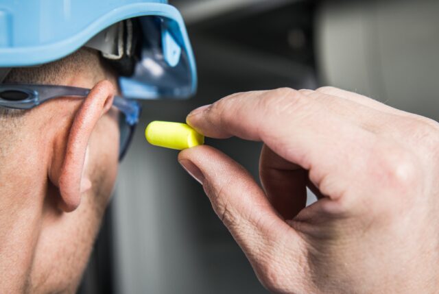 A person with a hard hat and safety goggles puts an earplug in his ear.