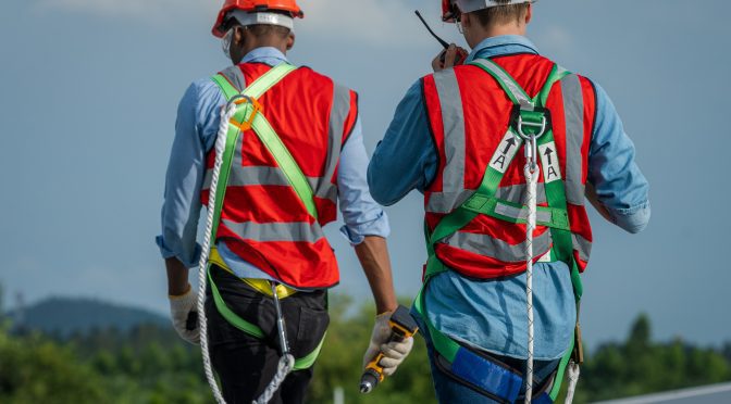 A pair of roofers in full PPE and safety equipment walk away from the camera, as one speaks into a radio