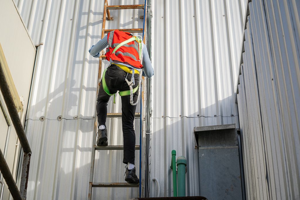 A roofer in a harness and safety gear ascends a ladder on the side of a commercial building