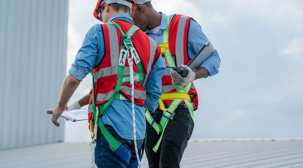 Two men in safety harnesses and vests discuss their commercial roof inspection checklist with their backs to the camera
