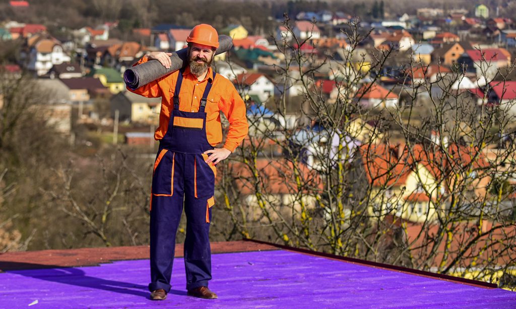 A roofer standing on a purple roof coating is in the process of installing BUR roofing on top of it. He wears purple overalls with an orange shirt and a hard hat.