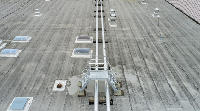 A view from above of a gray commercial roof bisected by two pipes running along the center with a short pair of stairs leading over them.